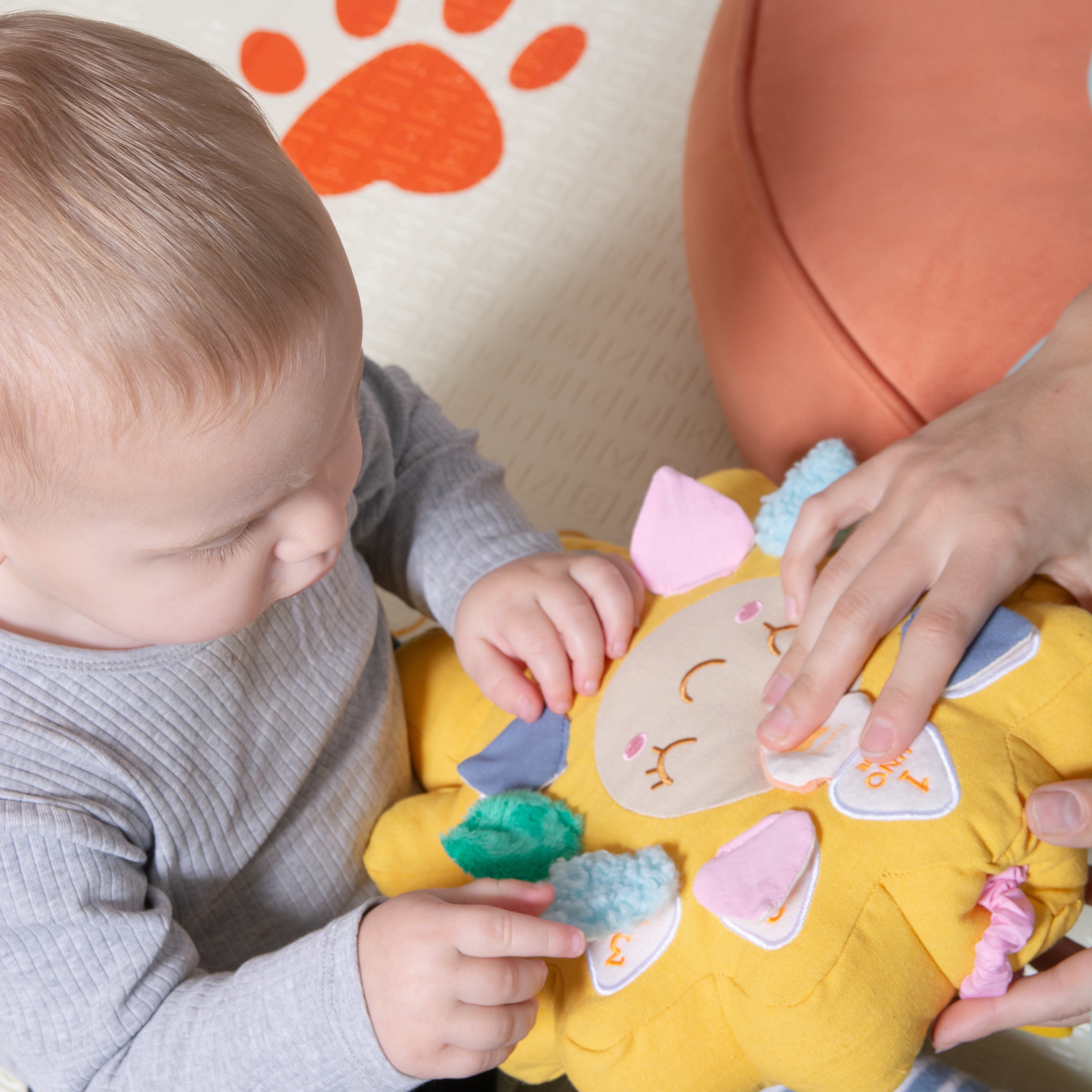 A baby in a gray ribbed long-sleeve shirt is sitting on a peach cushion and interacting with a yellow plush sensory toy with colorful fabric tabs, while an adult hand gently touches the toy and is showing him the number one