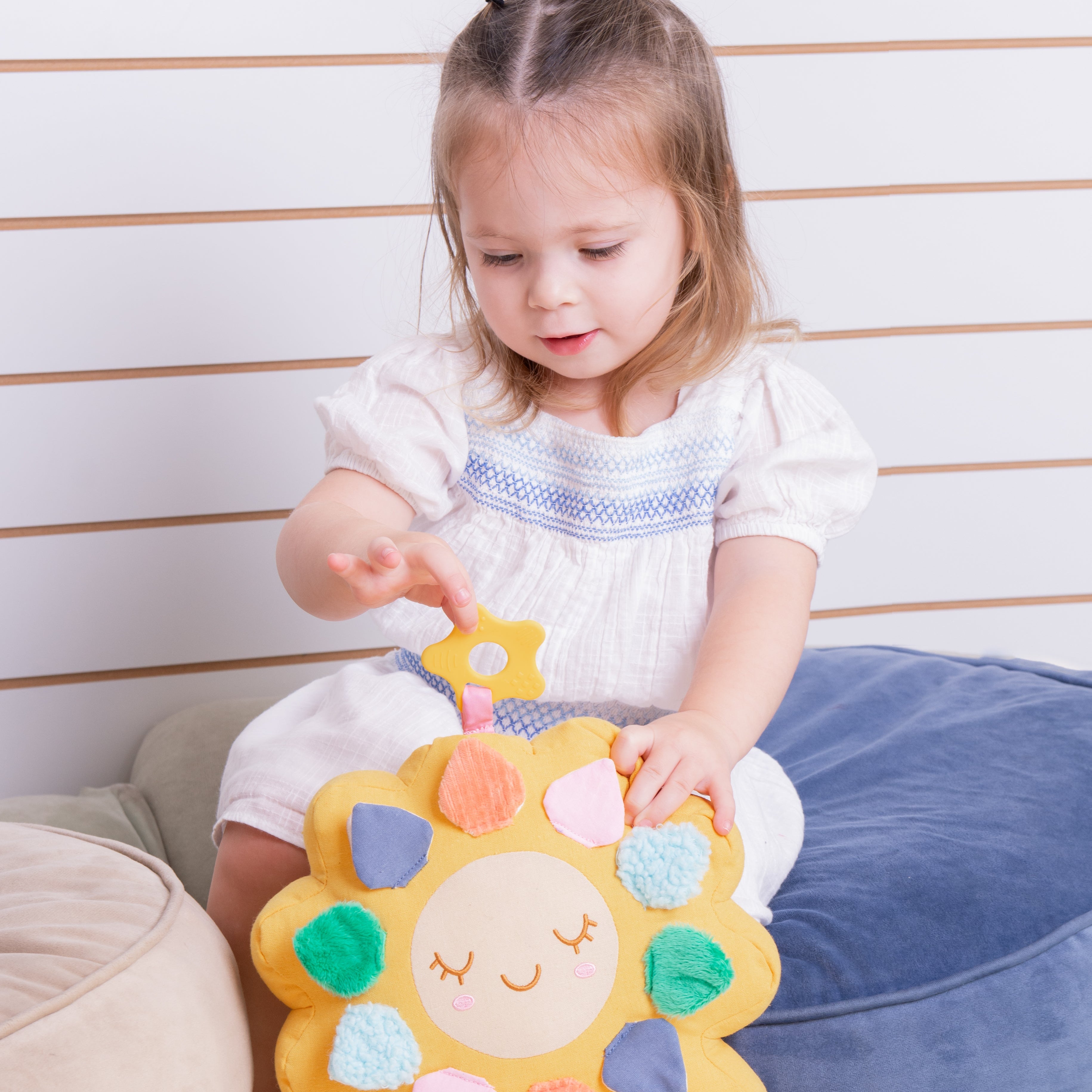 A young toddler girl in a white dress sits on a blue cushion, interacting with a yellow plush sun toy that has colorful fabric petals.