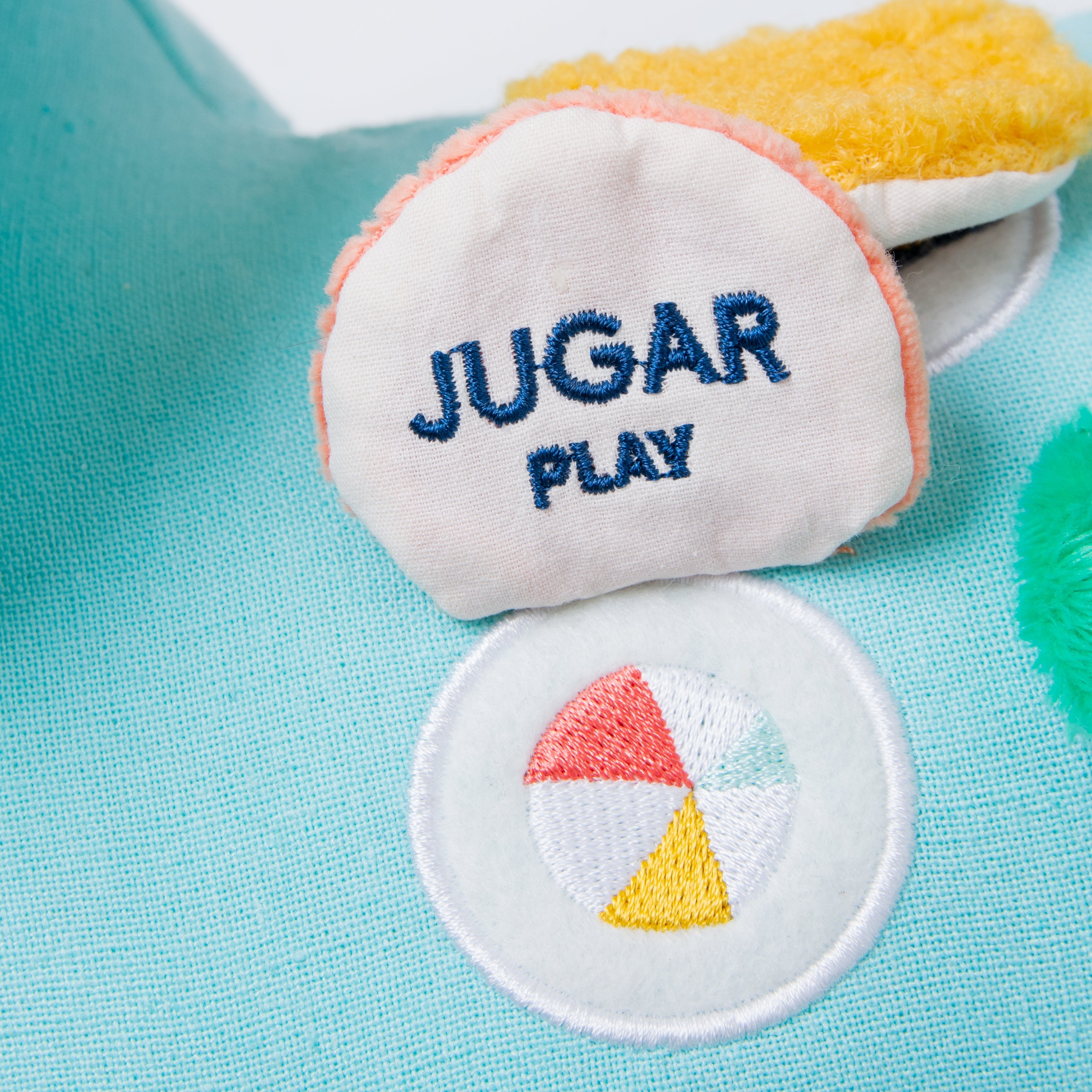 A close-up of a plush toy or cushion with embroidered text reading 'JUGAR PLAY' in blue on an off-white fabric surface, surrounded by pastel pink trim. Below it is a circular patch with a multicolor ball design in red, yellow, light blue, and white.
