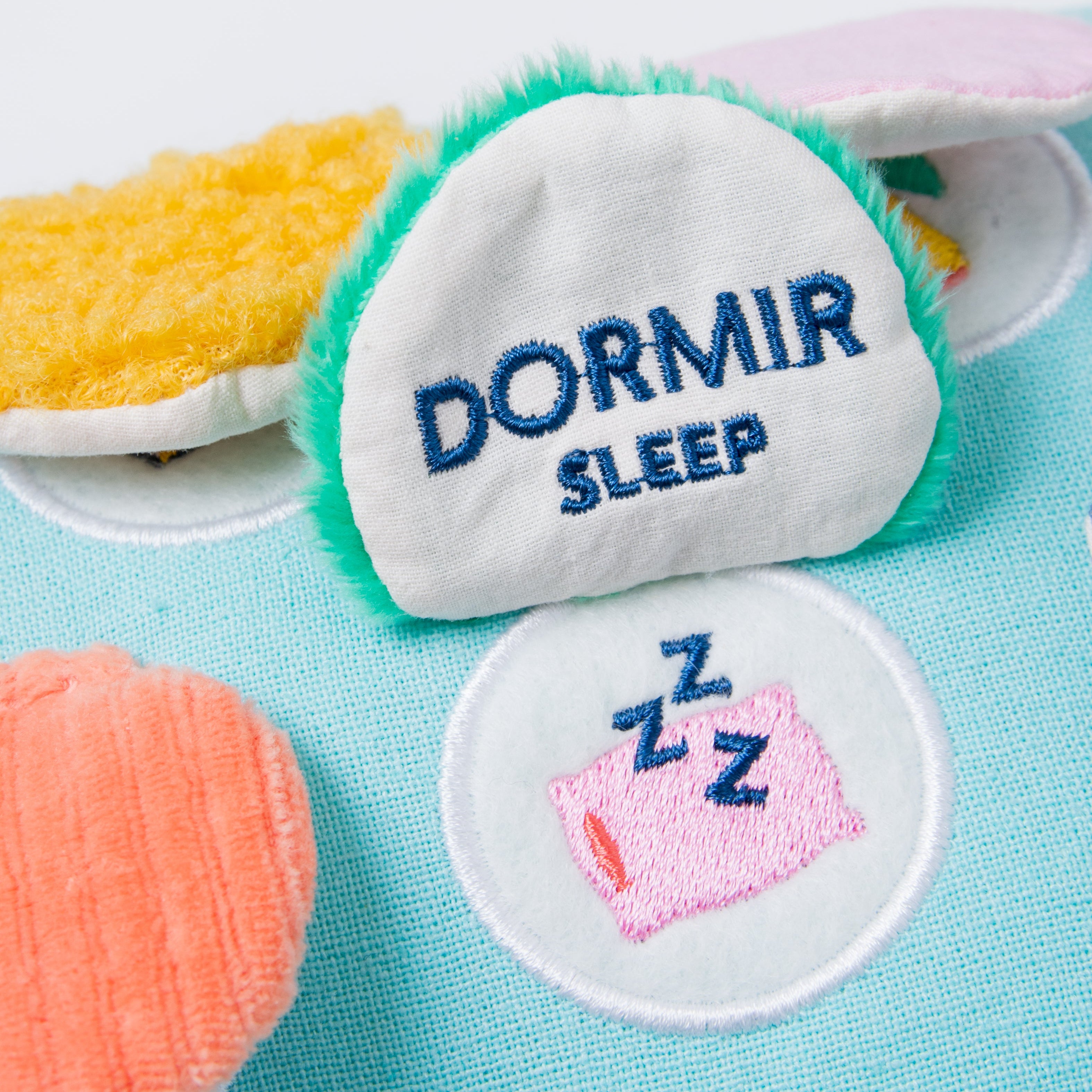 A close-up of a plush toy or comforter with embroidered patches. The main patch reads 'DORMIR SLEEP' in blue text on a white background with green faux-fur trim, while a smaller patch below shows a pink sleeping pillow with blue 'Z Z Z' letters.