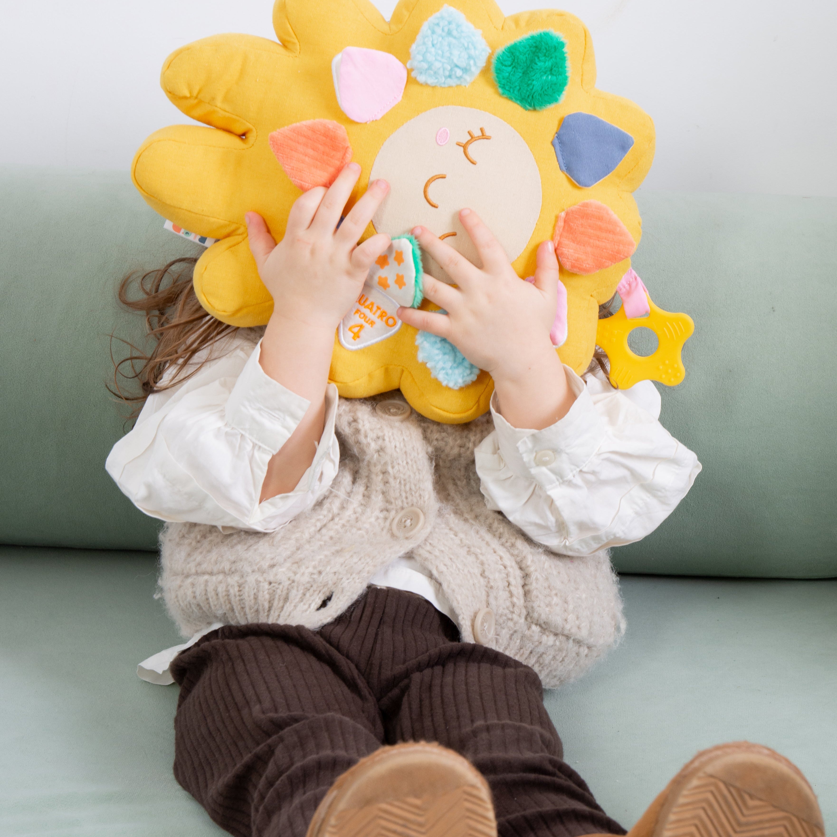 A child sitting on a mint green sofa, holding a large yellow sun-shaped plush pillow with colorful fabric petals and a small hanging toy in front of their face.