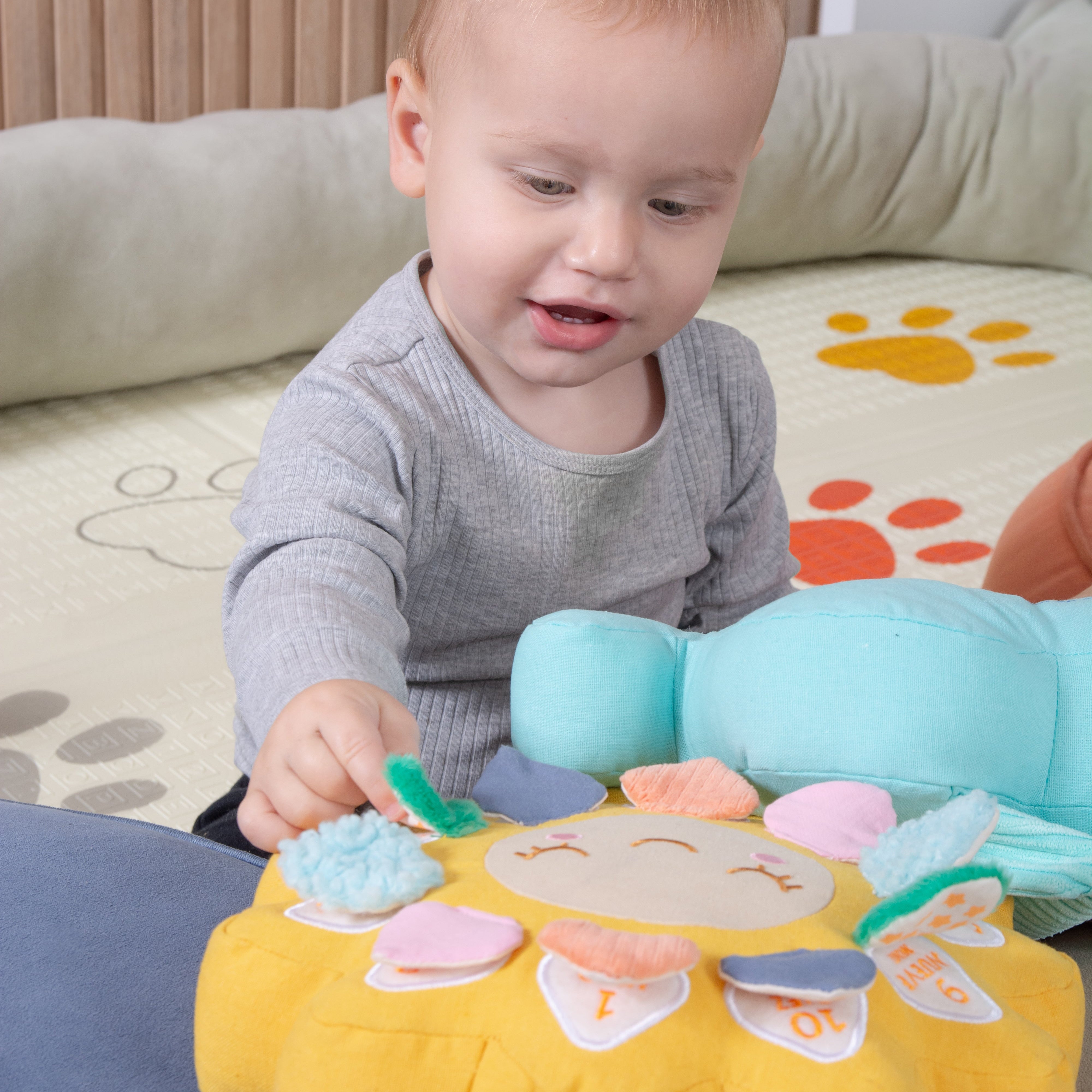 A baby sitting on a play mat, interacting with a colorful fabric sun toy and a light blue plush toy.
