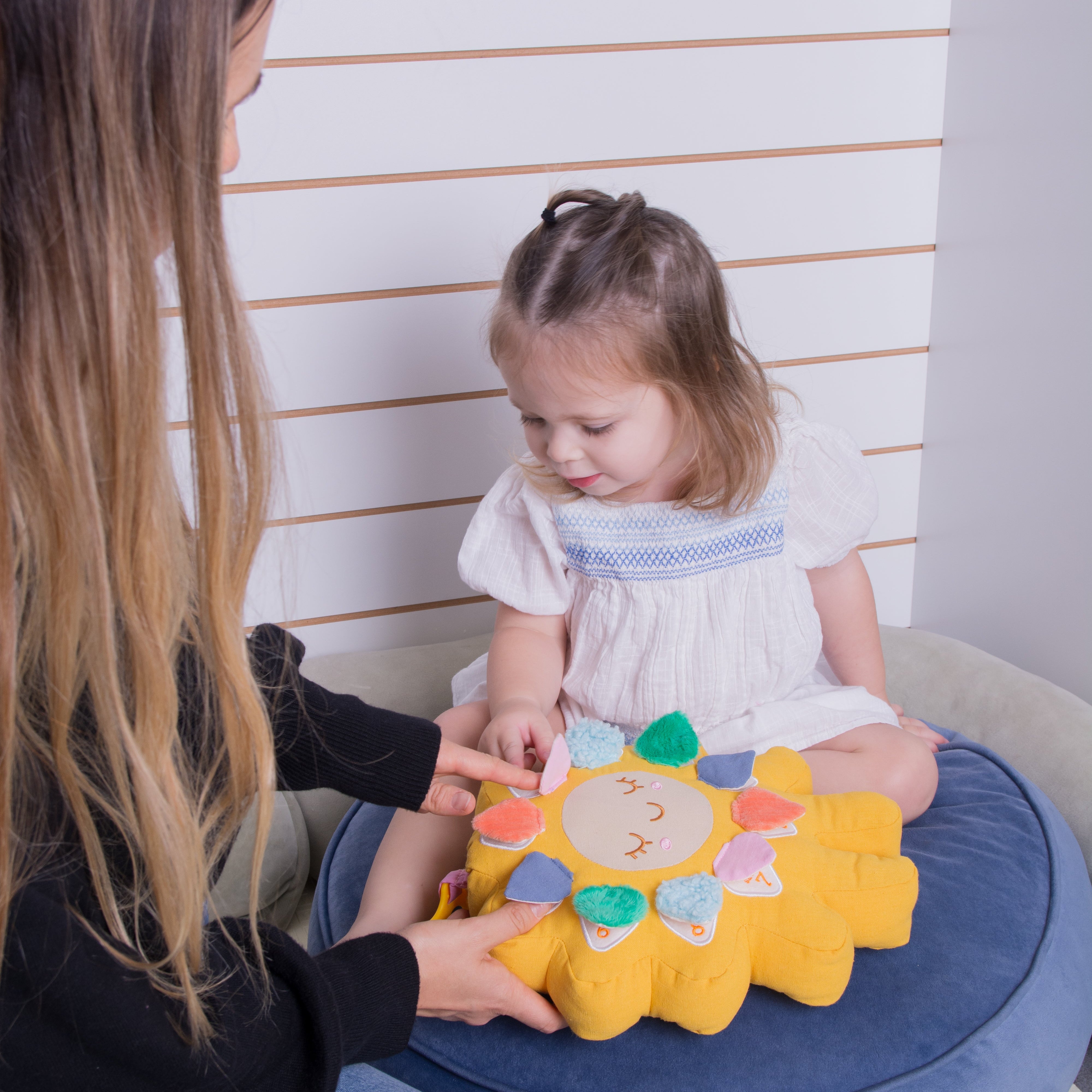 A young child sits on a round blue cushion while an adult points at a yellow sun-shaped sensory toy with colorful fabric petals. The setting is indoors against a white slatted wall.