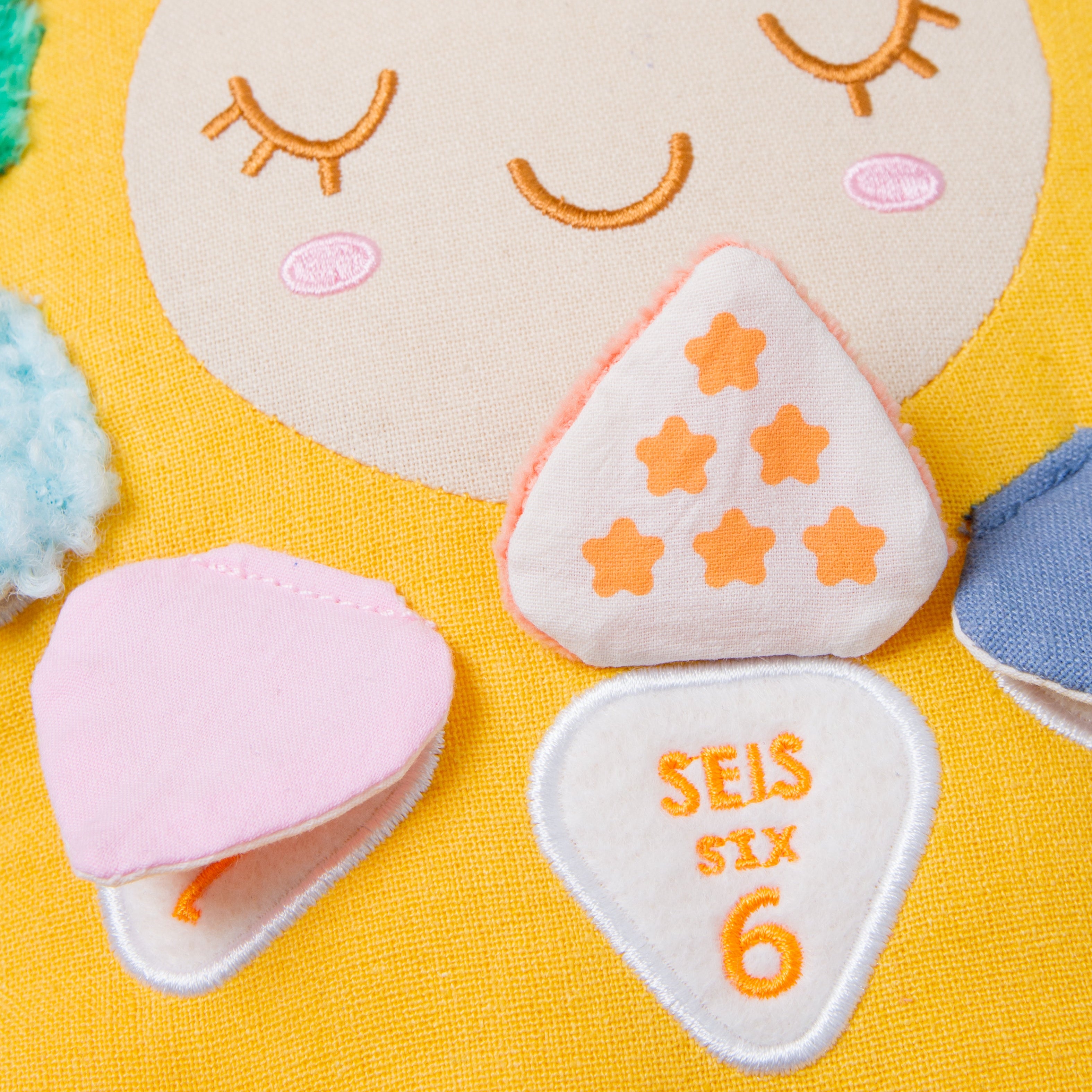 Close-up of a soft fabric sensory toy set on a bright yellow background, featuring a large beige face patch, a pink heart-shaped piece, a white triangle with orange stars, and a white teardrop-shaped piece with orange text reading 'SEIS SIX 6'.