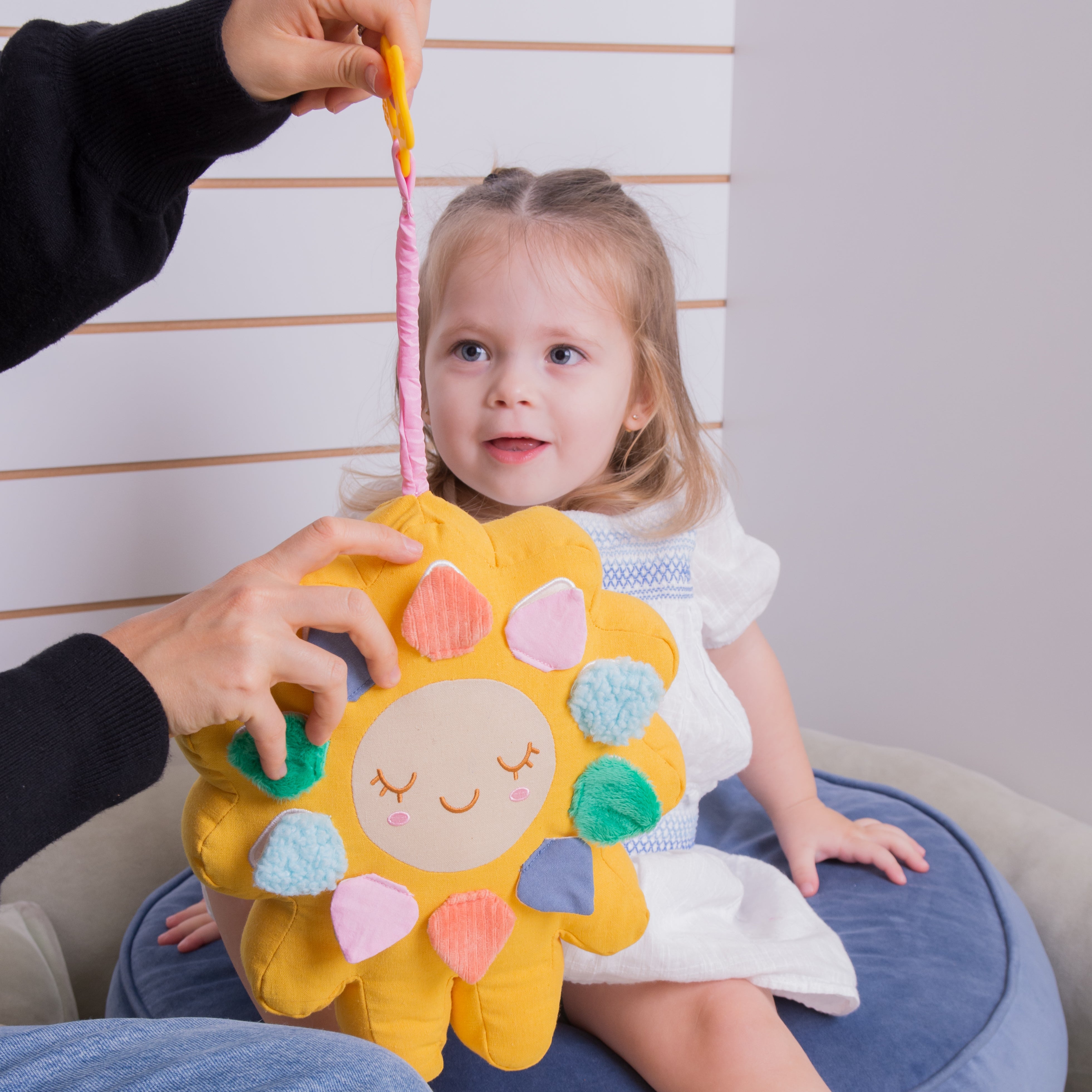 A young child sits on a blue cushion while an adult holds up a yellow sun-shaped plush toy with colorful fabric petals and a pink hanging loop. The scene is indoors against a light paneled. wall. The adult is pulling a music box string.