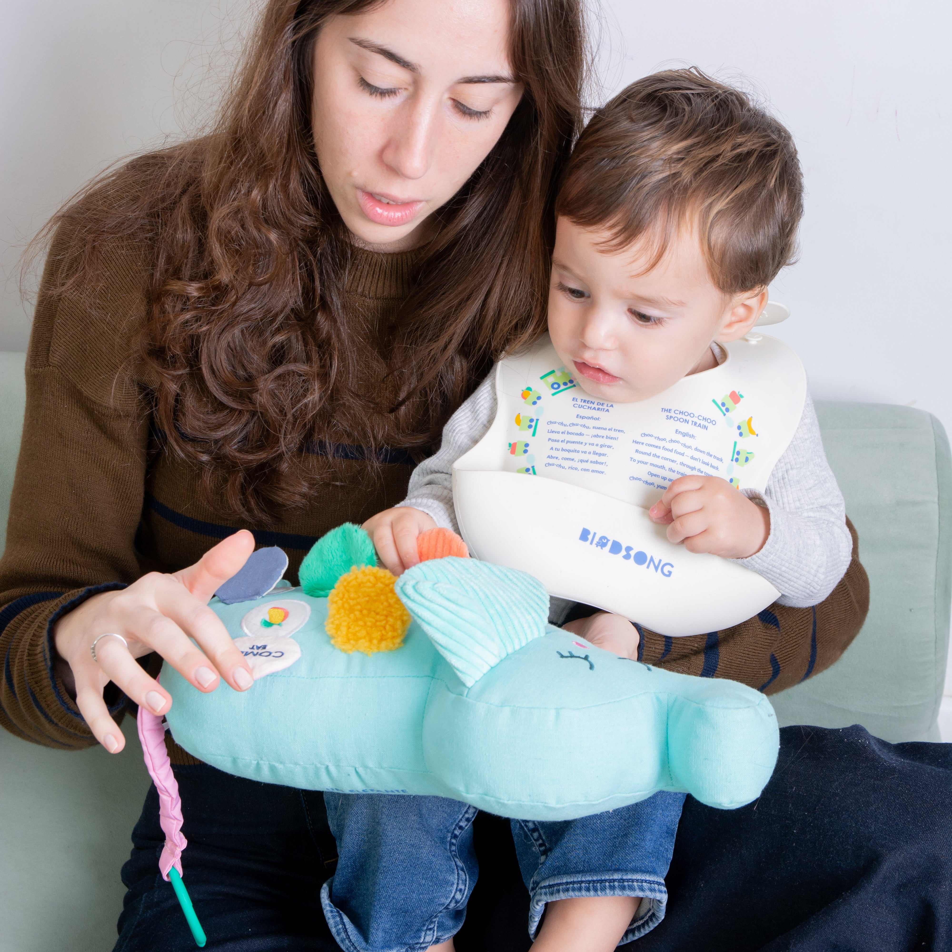 A woman and a baby are sitting together on a sofa, using a light blue plush baby bilingual toy with colorful sensory elements. The baby is wearing a white bib with the brand name 'BIRDSONG' visible.